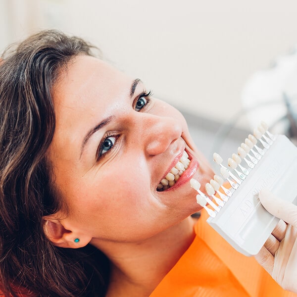 A woman at the dentist smiling as the doctor compares her teeth whiteness to a white color palette